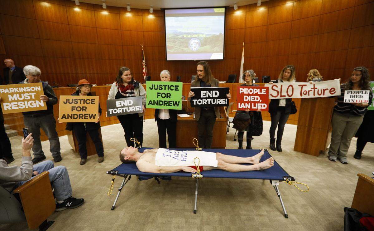 Protesters stand in San Luis Obispo County supervisors’ chambers in 2018 demanding justice for the death of Andrew Holland, who died in County Jail custody after 46 hours in a restraint chair.
