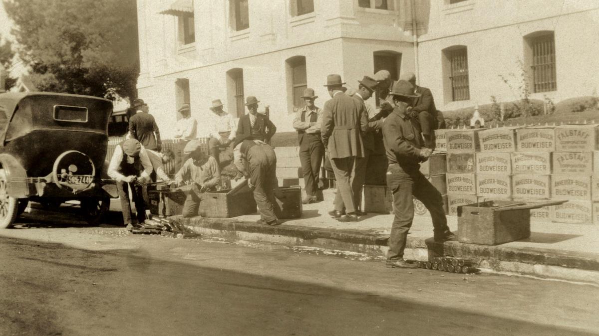 San Luis Obispo County Sheriff’s deputies dispose of illegal beer and Canadian whiskey in front of the courthouse, after a prohibition raid circa 1921. Crates labeled Falstaff and Anheuser-Busch are on sidewalk as the gutter fills with suds.