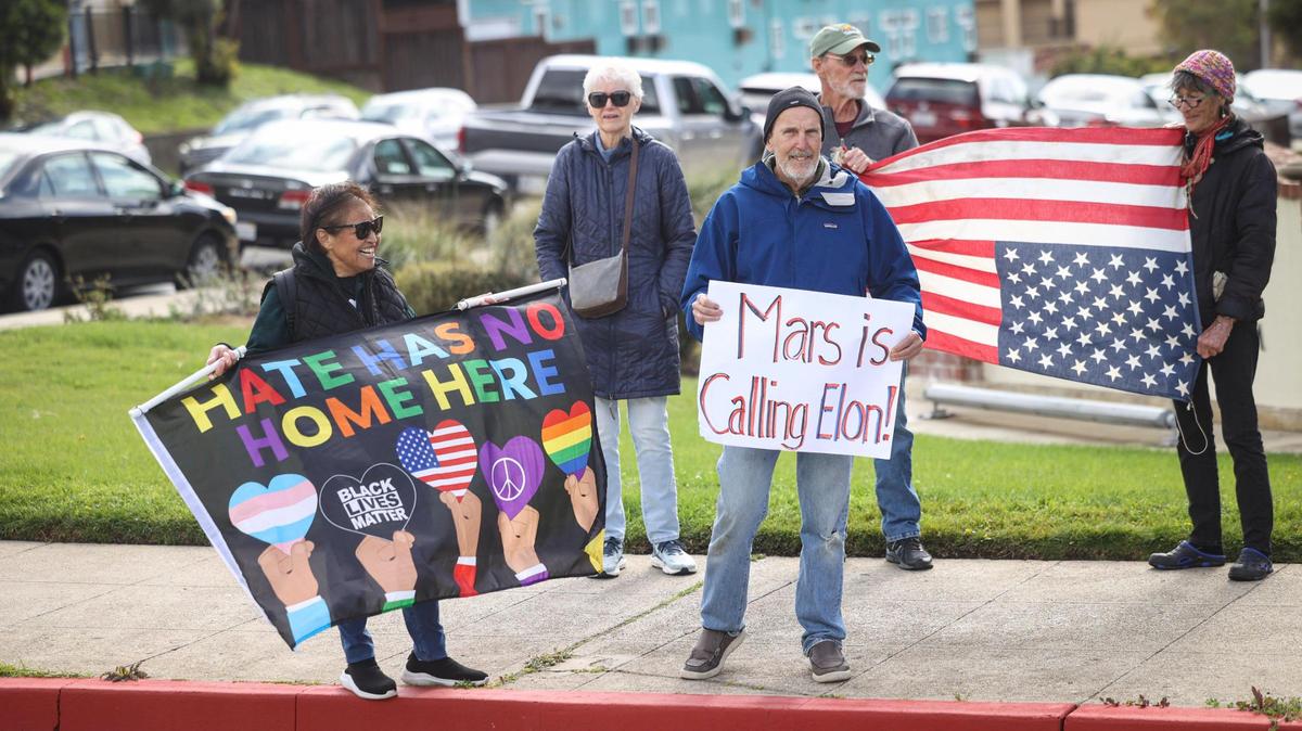 Jeanie McCombs and Rick Wolfe, front, hold signs on Grand Avenue during a rally in support of veterans. Over 50 protesters gathered near the Veterans Memorial Building in San Luis Obispo with signs in support of veterans services and critical of President Trump and Elon Musk on March 14, 2025.