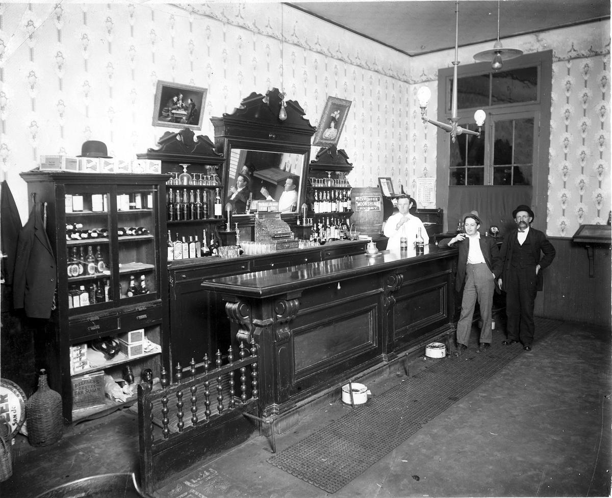 An circa 1910s photo of an unidentified San Luis Obispo saloon shows cigars labeled San Luis Perfectos under a bowler hat at the left. A bottle cap calendar on the wall shows this was a day in a leap year February. Gas lamps are lit, while electric lamps are not.