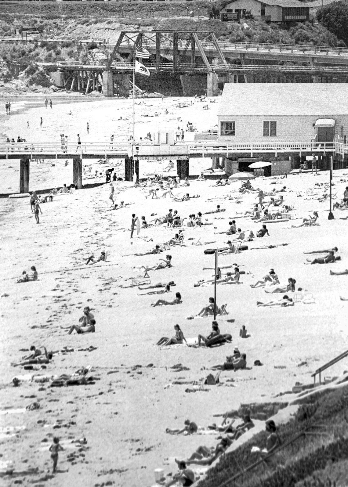 Beachgoers sit in sun with old Pacific Coast Railway bridge in background. Views of Avila Beach on June 28, 1976.