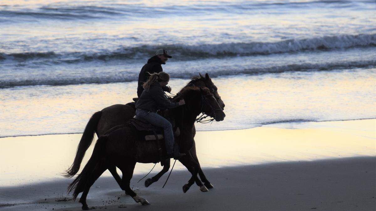 Two people ride horses along the beach.