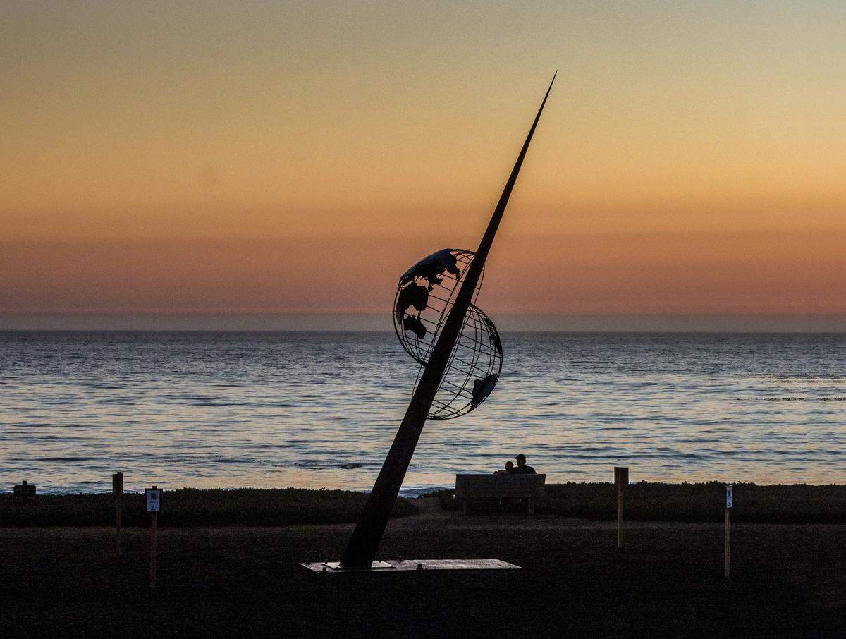 As the sun sets on Oct. 1, 2021, at a San Simeon shoreline, a couple cuddles on a bench near “Shift,” a new 1,200-pound sculpture.