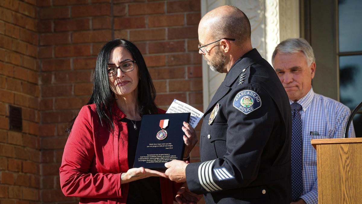 El jefe de policía de Atascadero, Dan Suttles, entrega un premio por salvar vidas a María Contreras, tía de José Gutiérrez, quien fue atropellado y asesinado por un camión que salió disparado de El Camino Real el 15 de marzo de 2025. José, quien siempre quiso ser policía, le advirtió a un amigo que se apartara antes de que lo atropellaran. Contreras habló y aceptó el premio en nombre de la familia en una ceremonia frente al Ayuntamiento el 22 de abril de 2025. A la derecha, el alcalde Charles Bourbeau.