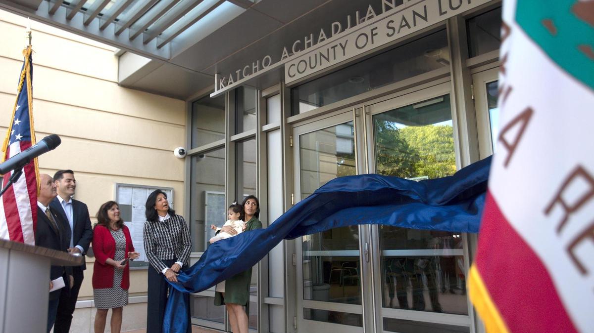 The new Katcho Achadjian Government Center sign was unveiled in a ceremony Friday, Oct. 20, 2023, in front of the former San Luis Obispo County Government Center. SLO County Supervisor John Peschong emceed the festivities. Achadjian’s wife Araxie, his daughter Nyri and son Hratch all spoke at the podium before Araxie pulled down a blue cloth revealing the new sign. From left, SLO County Supervisors John Peschong, Jimmy Paulding and Debbie Arnold, watch as Araxie unveils the sign.