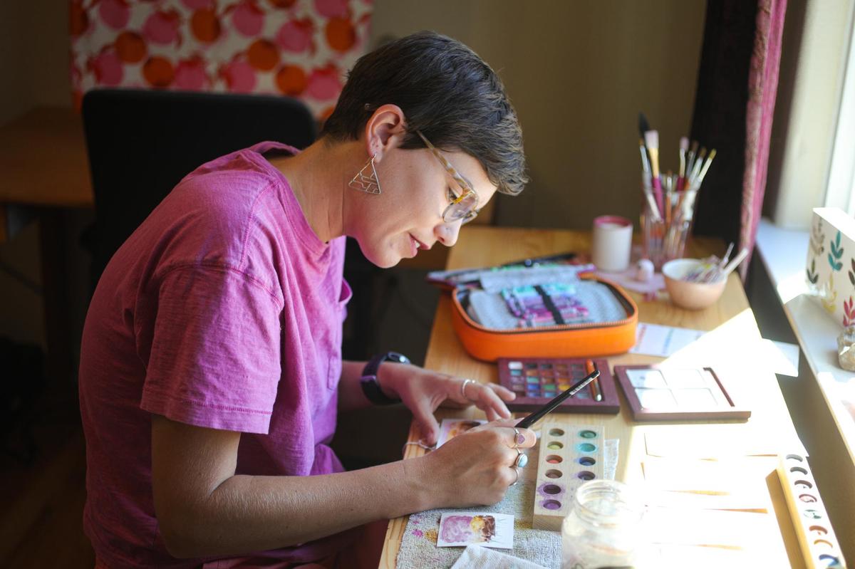 person in a pink shirt sitting at a desk over a piece of paper and a tray of paints. 