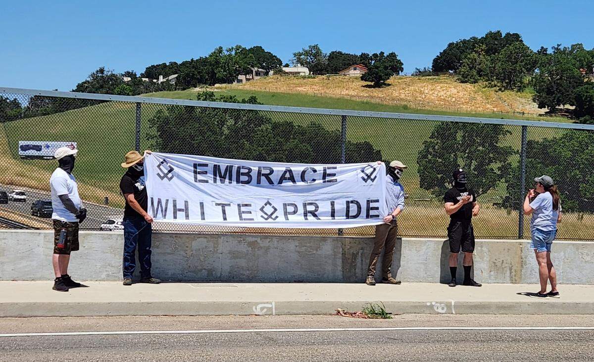 Several men with masks hold up an “Embrace white pride” flag on the Vineyard Drive Highway 101 overpass on Saturday, May 13, 2023.