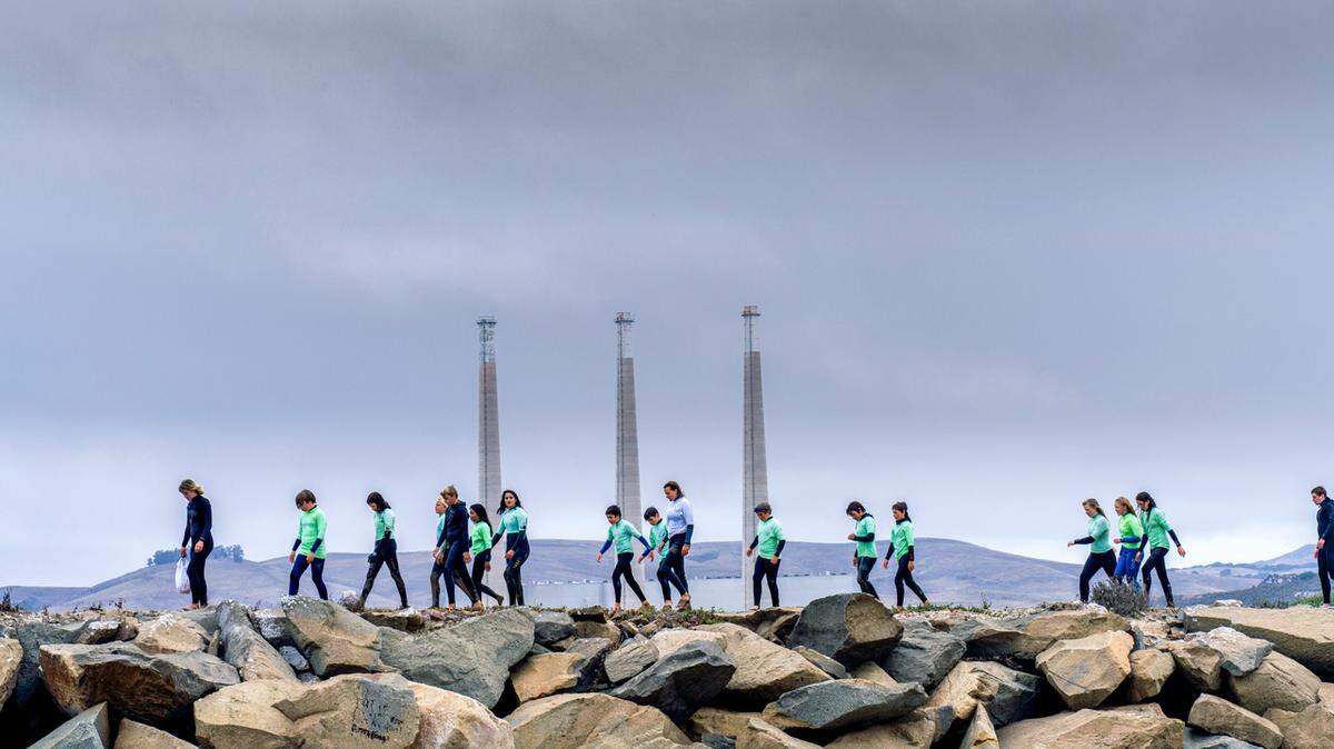 Junior Lifeguards participants walk along the sand spit in Morro Bay with the stacks of the former power plant towering behind them in July 2020. The city is hosting a Zoom forum Wednesday, Sept. 8, to discuss possibly removing the stacks.