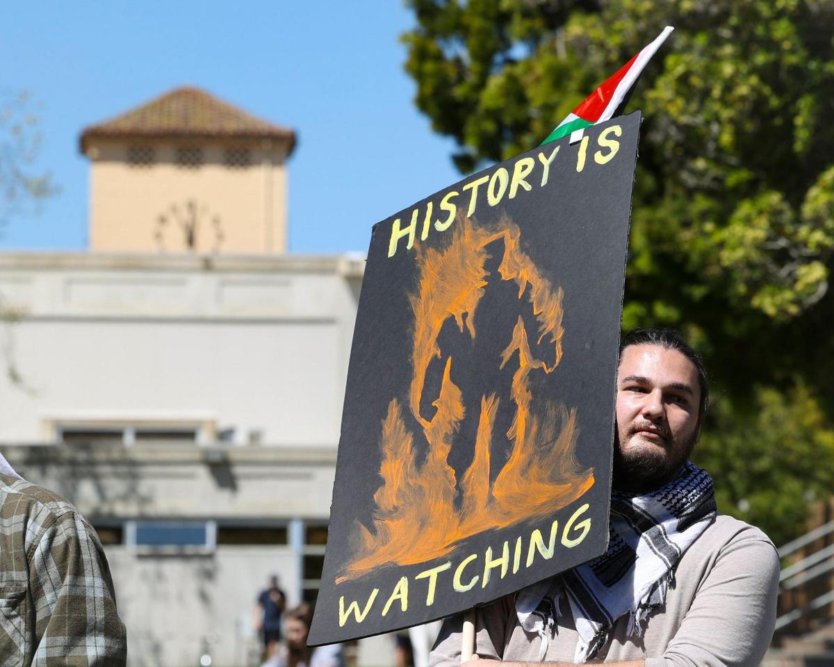 Brooks Martin, a Cal Poly grad student, holds a sign that reads “History is watching” as about 70 people gathered on Cal Poly’s Dexter Lawn on May 1, 2024, to stage a silent die-in in support of Palestine.
