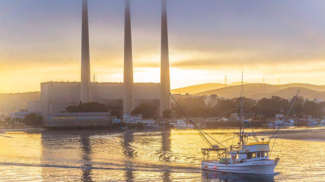 The Morro Bay-based Michael Too heads out to sea on a salmon-fishing trip as the sun rises behind the shuttered Morro Bay Power Plant in July 2019. The city of Morro Bay told Vistra Corp. that it must remove the power plant’s iconic stacks by 2028 or pay a hefty fine.