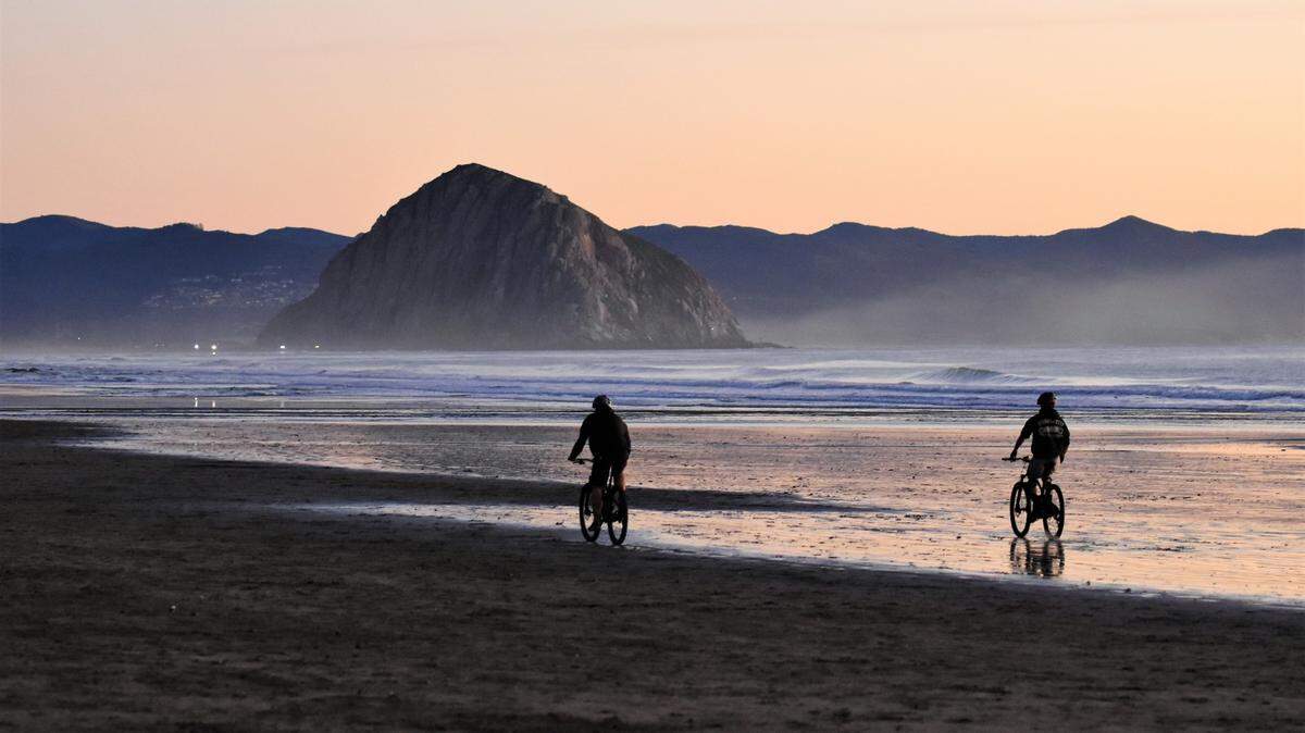 Bike riders on the beach are heading toward Morro Rock during low tide.