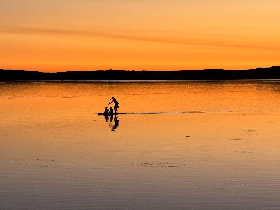 A paddleboarder with two kids crosses Morro Bay at sunset.