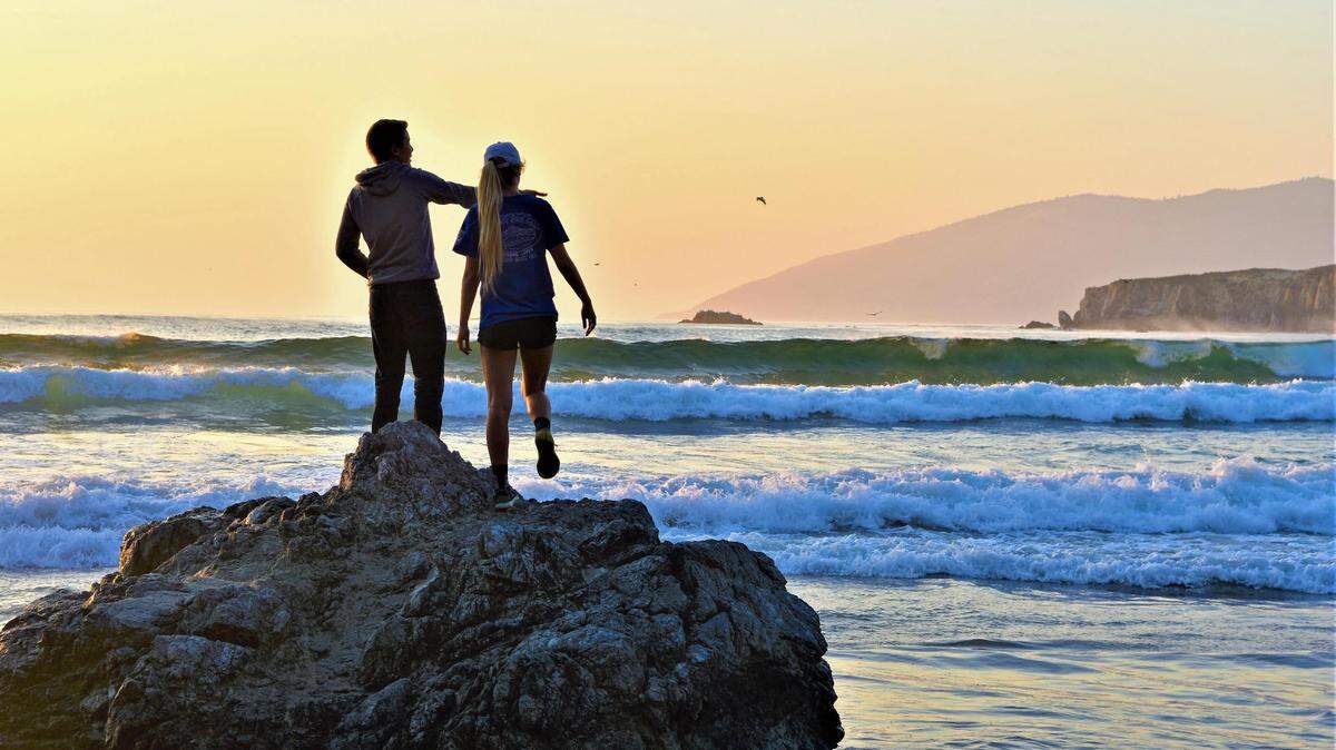 Two people watch waves crash along the Big Sur coastline.