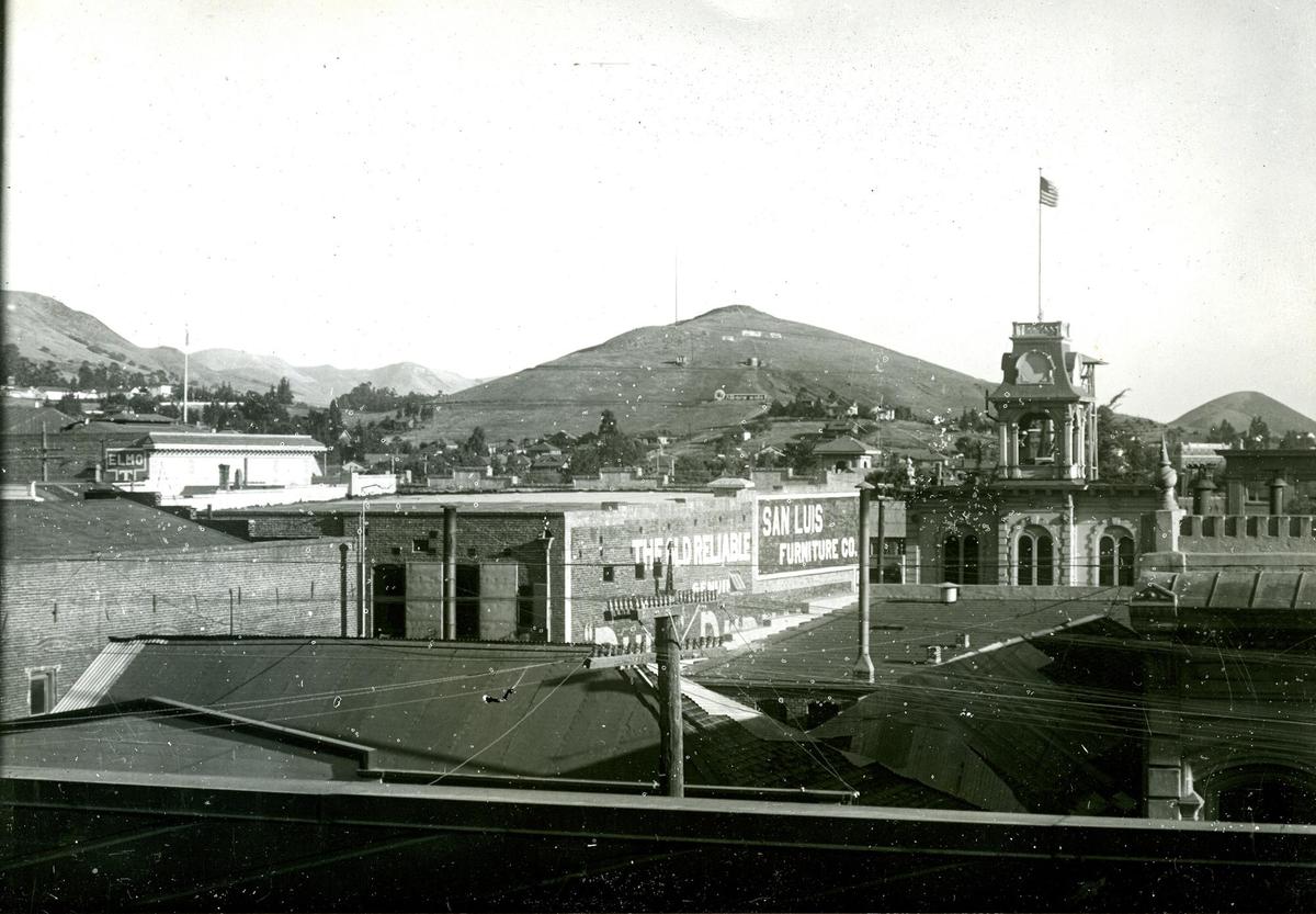 A photo of downtown San Luis Obispo shows Terrace Hill before it had the top removed. Buildings that are now gone are the City Hall at right with flag pole and Elmo Theater at left. Sinsheimer building is at corner lower right. Estimated year time frame is 1920s-30s.