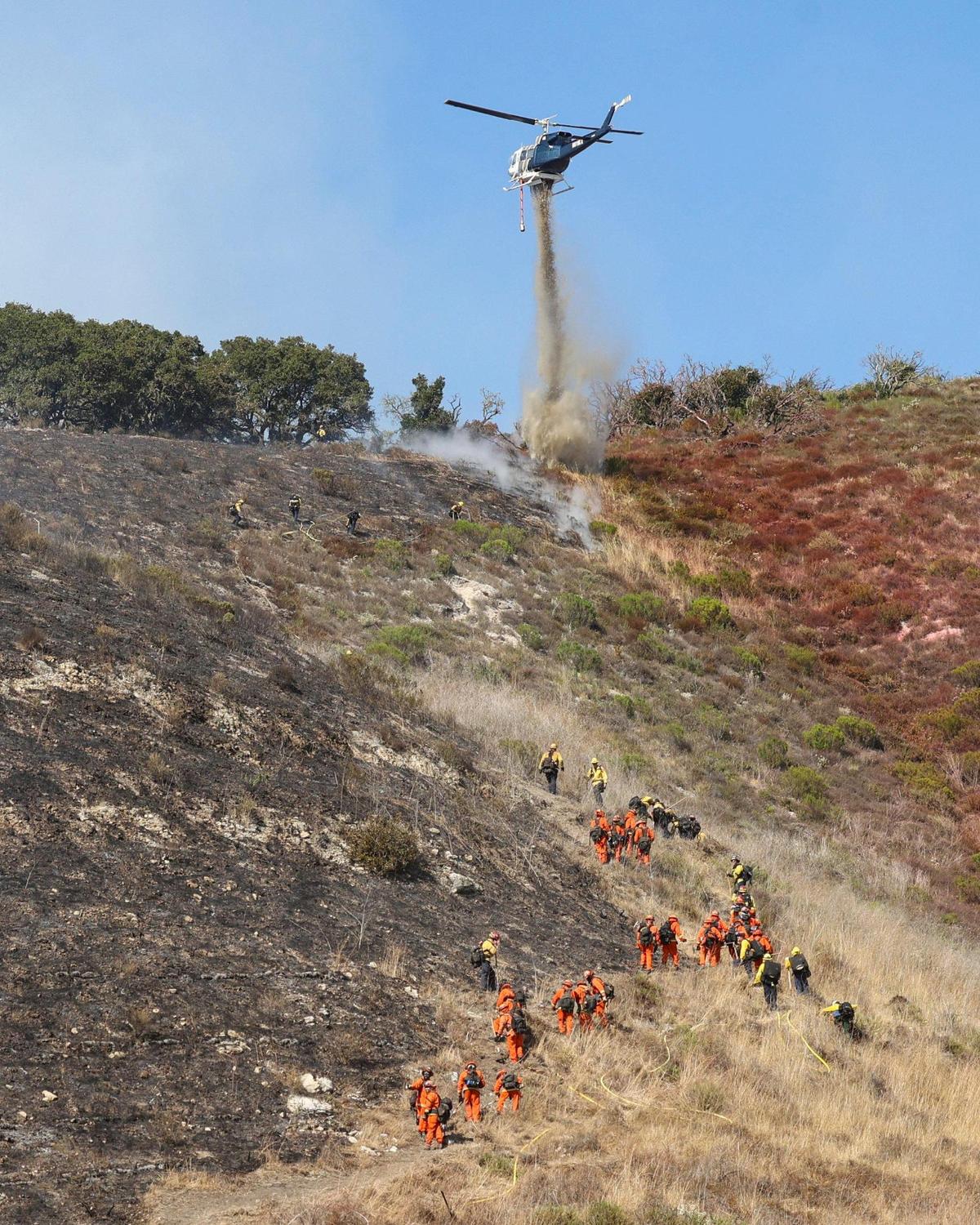 Helicopter works on a ridge line above hand crews cutting fireline. Firefighters responded to a wildland blaze on the edge of Pismo Beach on June 9, 2025.
