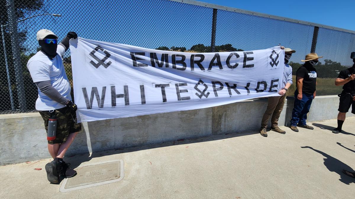 Several men with masks hold up an “Embrace white pride” flag on the Vineyard Drive Highway 101 overpass on Saturday, May 13, 2023.