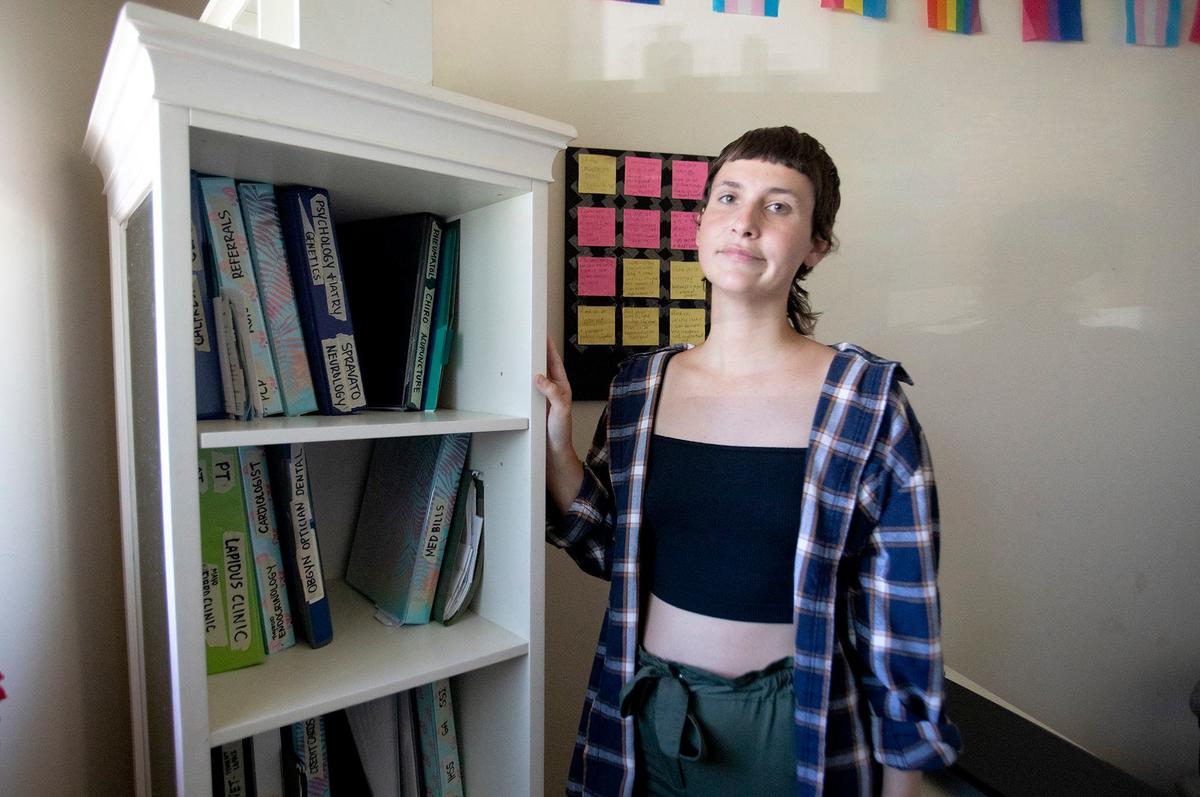 Faith LeGrande, a San Luis Obispo resident who suffers from multiple dynamic disabilities, is working to help connect others with resources through their business Art and Soul SLO. Legrande stands next to a bookshelf filled with binders of their health records.