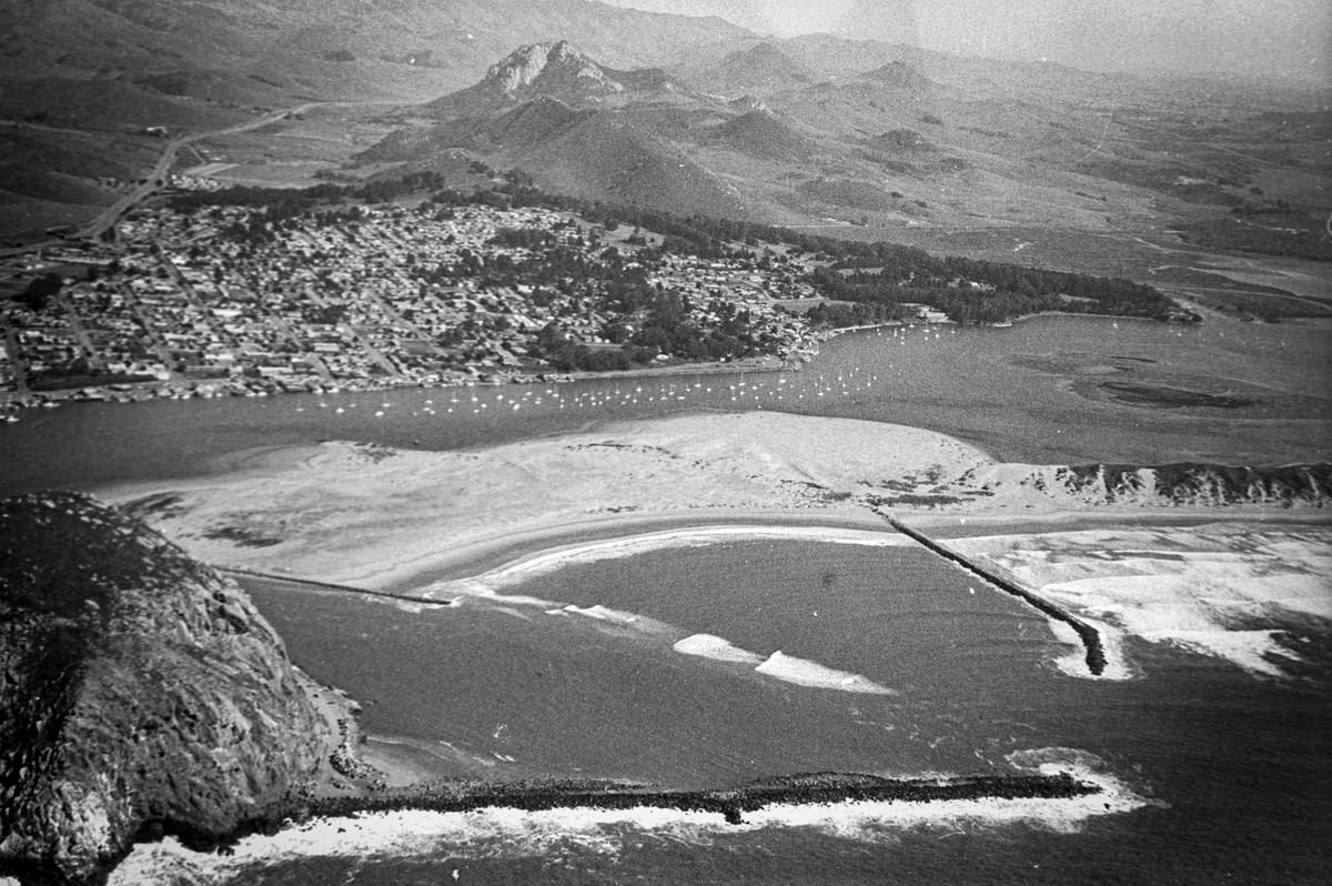 Morro Bay harbor mouth in this historic aerial view shows waves breaking due to sand build up. A year earlier the San Mateo sank with 23 school children aboard. Two adults died as a result of the accident. Heroic efforts of the Harbor Patrol and others rescued most. Highway 1 and the morros are in the background. Reporter Brooks Townes took a flight over coastal areas from Morro Bay to Avila Beach on Feb. 22, 1984.