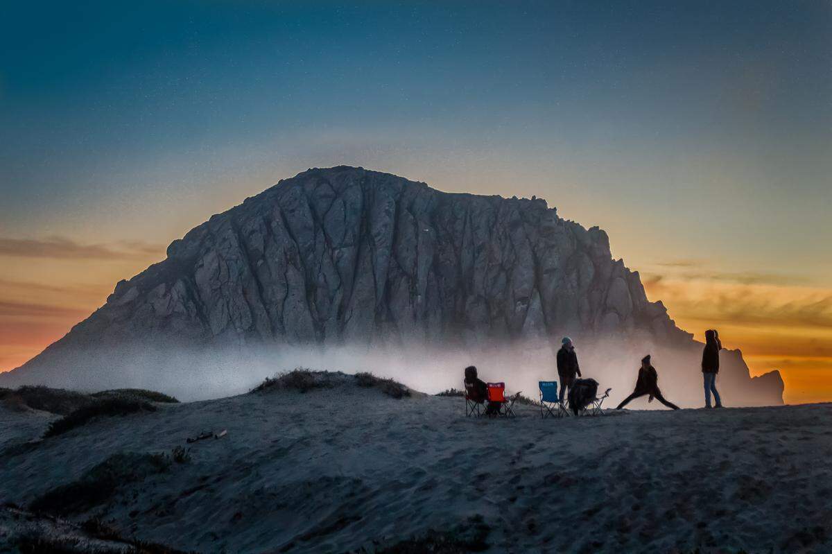 Bundled-up beach-goers are silhouetted against a low ribbon of fog during a recent sunset at Morro Rock.