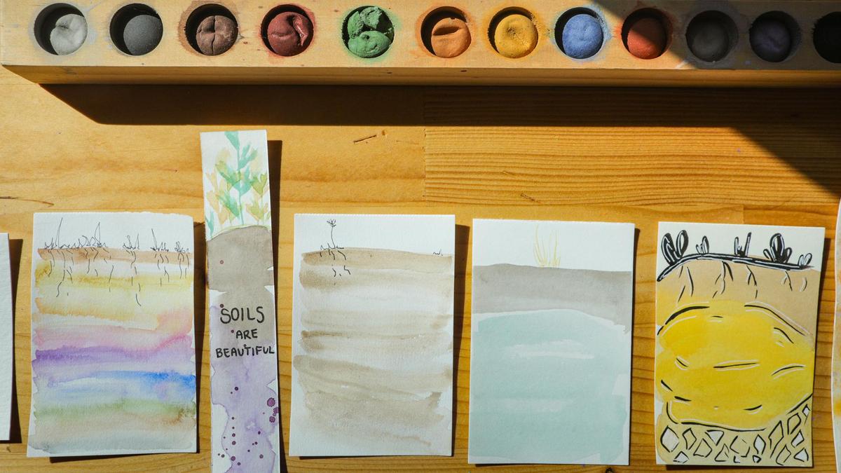 Several paintings laying side by side on a desk next to a tray of water color paints