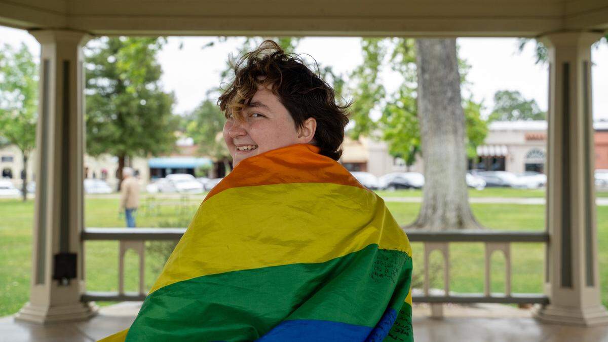 Paso Robles High School Queer Student Union president Aster Watson poses for a photo while wearing a Pride flag at Downtown City Park in Paso Robles on Saturday, May 2, 2026. The flag is covered in signatures and messages of Queer Student Union members.