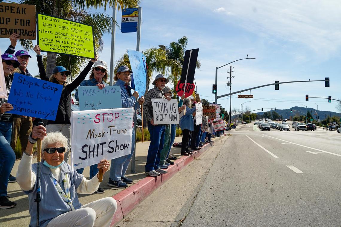 Protesters hold signs and chant to show their opposition to the policies of Donald Trump’s second term in office and Elon Musk’s Department of Governmental Efficiency, on Los Osos Valley Road on Monday, Feb. 17, 2025.