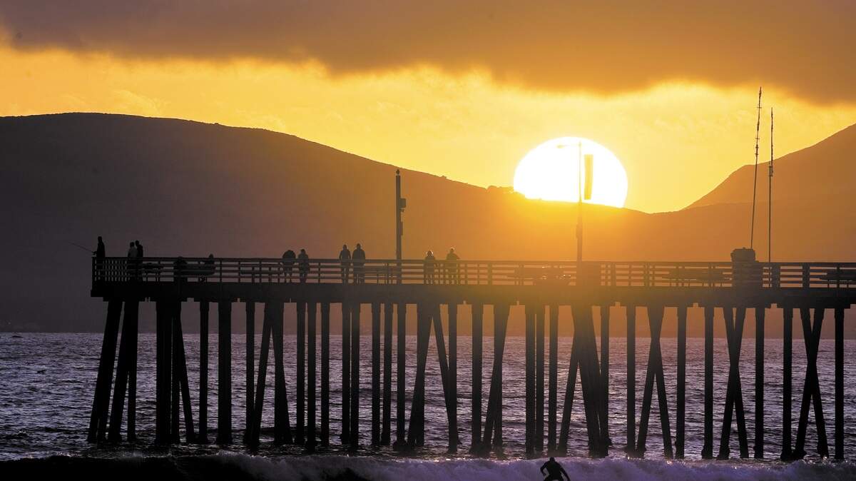 A band of fog stretches above the setting sun as a surfer catches a wave near the Pismo Beach Pier.