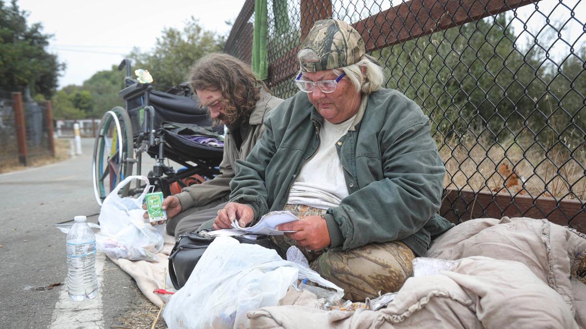 Nena Kosbab-Jones looks through medical paperwork as partner Matthew Jones has a snack. She uses the wheelchair and he said he had been bruised during a recent arrest and was just out of jail. He has a snack before taking the bus to recover confiscated personal items like their phones. They were on the Bob Jones Trail near San Luis Creek, a short distance from 40 Prado Homeless Services Center, on Oct. 9, 2024.