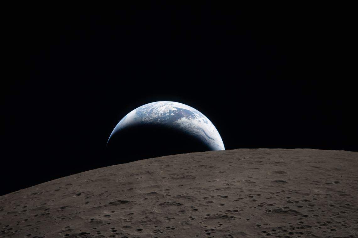 Earthset captured through the Orion spacecraft window at 6:41 p.m. EDT, April 6, 2026, during the Artemis II crew’s flyby of the Moon. A muted blue Earth with bright white clouds sets behind the cratered lunar surface. The dark portion of Earth is experiencing nighttime. On Earth’s day side, swirling clouds are visible over the Australia and Oceania region. In the foreground, Ohm crater has terraced edges and a flat floor interrupted by central peaks—formed when the surface rebounded upward during the impact that created the crater.