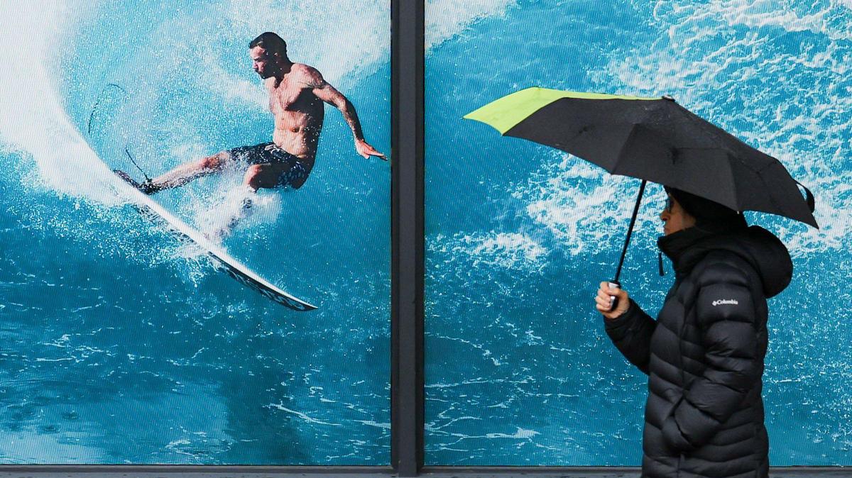 A pedestrian walks beneath an umbrella beside a surfing display in the window of Moondoggies in downtown San Luis Obispo on March 9, 2023, as a new atmospheric river storm hit the Central Coast.