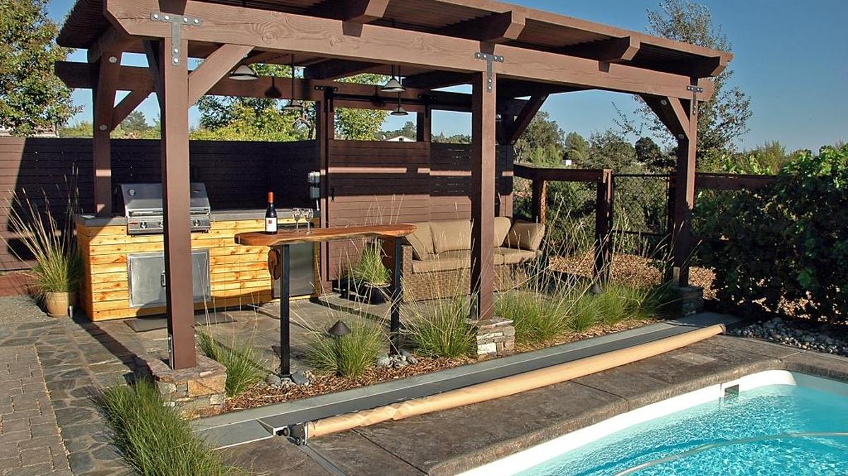 The new outdoor kitchen is surfaced in flagstone with brown grout to match the existing pool deck. The bar, with Slender Veldt Grass, Pennisetum spathiolatum, below, was handcrafted from a recycled cypress log. Horizontal siding covers the existing fence and continues on a sliding barn door to screen pool equipment.
