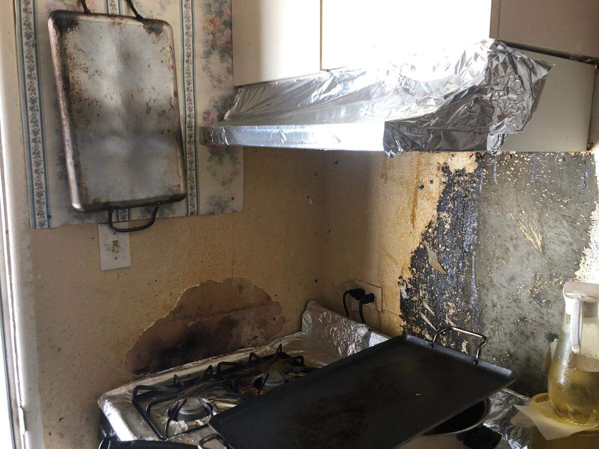A stove in a Grand View Apartments kitchen pressed up against a wall, causing it to become a fire hazard due to cooking residue. The area behind the stove is also in need of repairs.