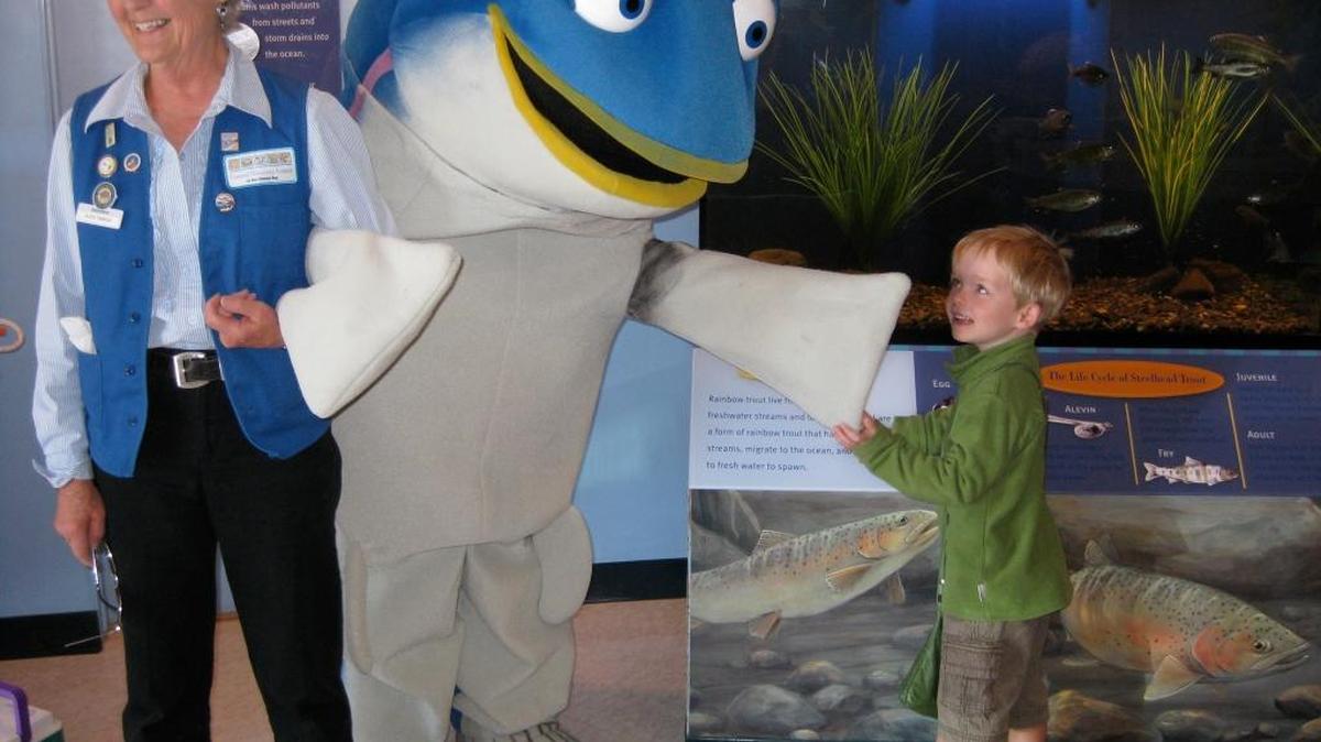 Leslie McGarry, in costume as Sammy Steelhead, joins a Coastal Discovery Center docent, Alice Yamada, in greeting a visitor.