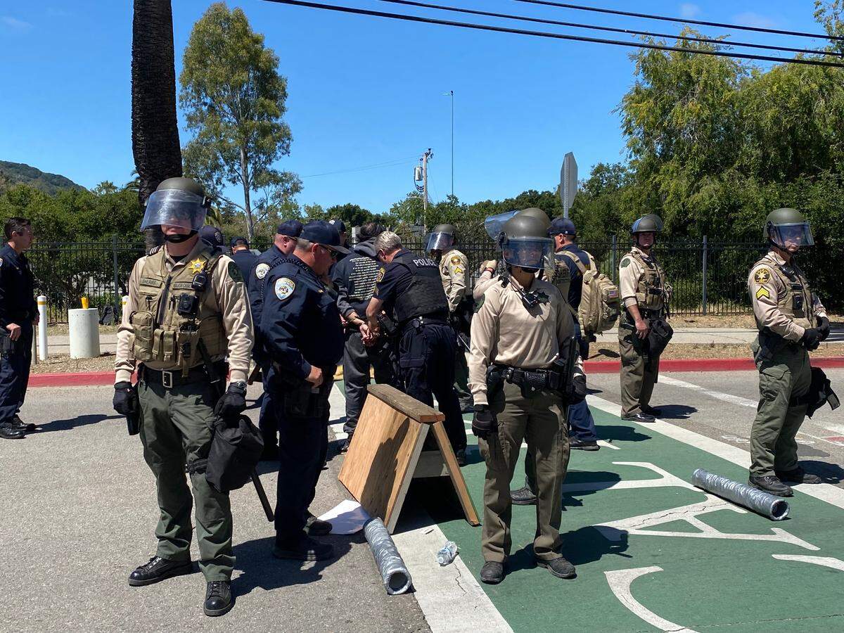 Police detain a pro-Palestine protester who chained himself to a barricade blocking the California Boulevard and Campus Way intersection near Cal Poly on May 23, 2024, while sheriff’s deputies in riot helmets form a perimeter.