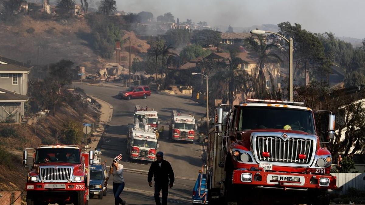 People walk by parked fire trucks in a residential area where a wildfire destroyed several homes Dec. 5, 2017, in Ventura.