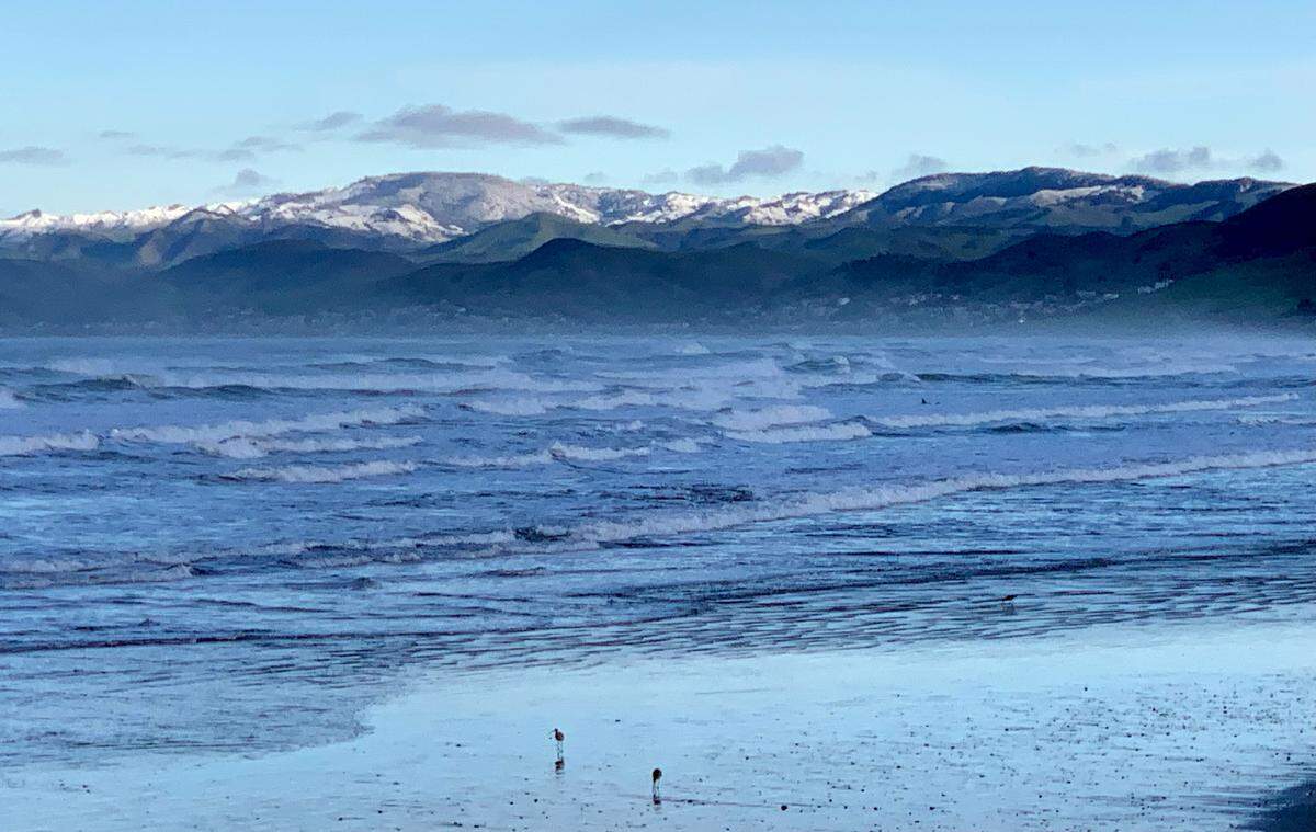 Snow dusts the Santa Lucia Mountains behind Cayucos in a view from the beach at Morro Rock on Thursday, Feb. 23, 2023.