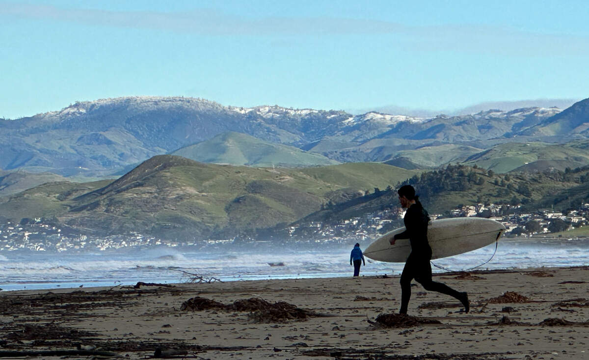 A surfer heads to the waves in Morro Bay in front of a snowy mountains on Saturday, Feb. 25, 2023.