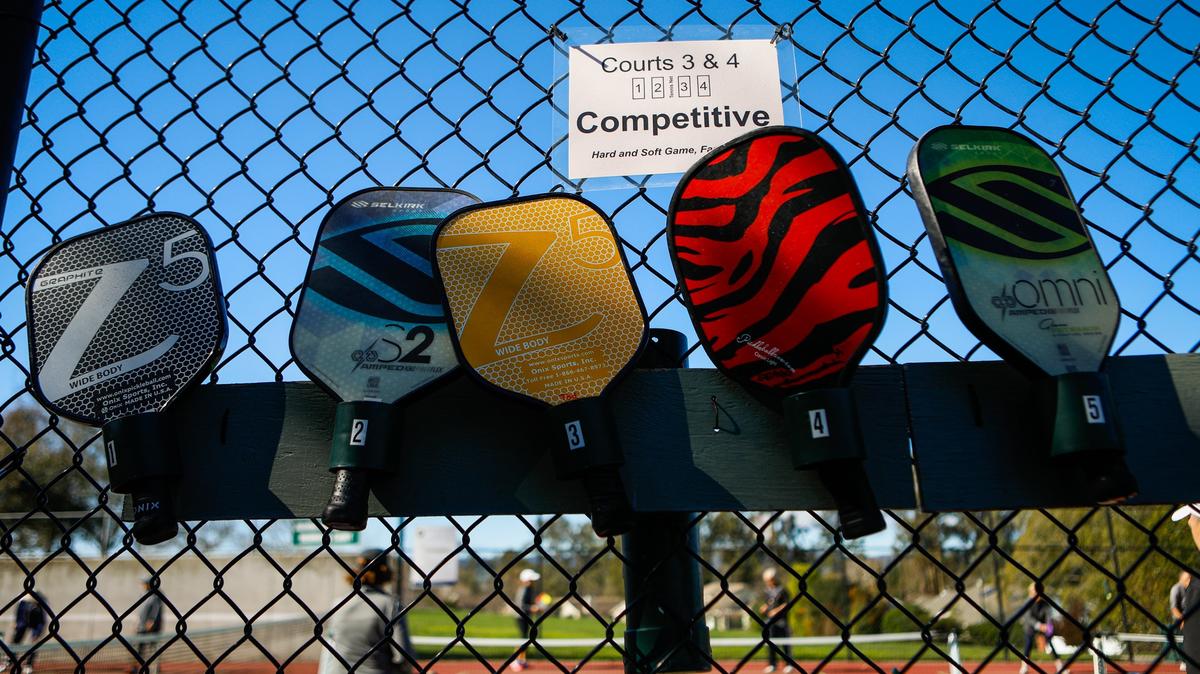 Pickleball paddles hang on a fence by those awaiting an open court at E A French Park in San Luis Obispo.