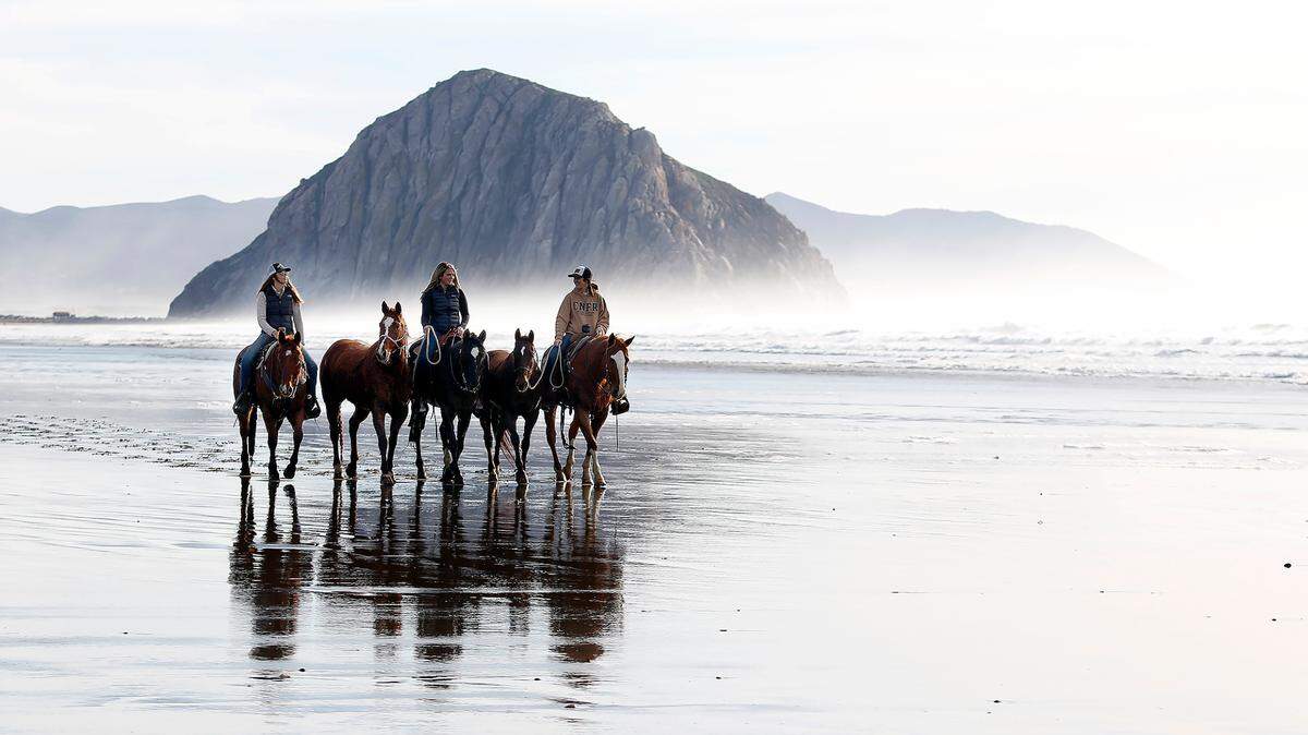 Framed by Morro Rock, Cal Poly students Lyndsey Goldwyn, left, Sydney Goldwyn and Taylor Marrou exercise their horses on the beach during a break in wet weather on Jan., 6, 2023.