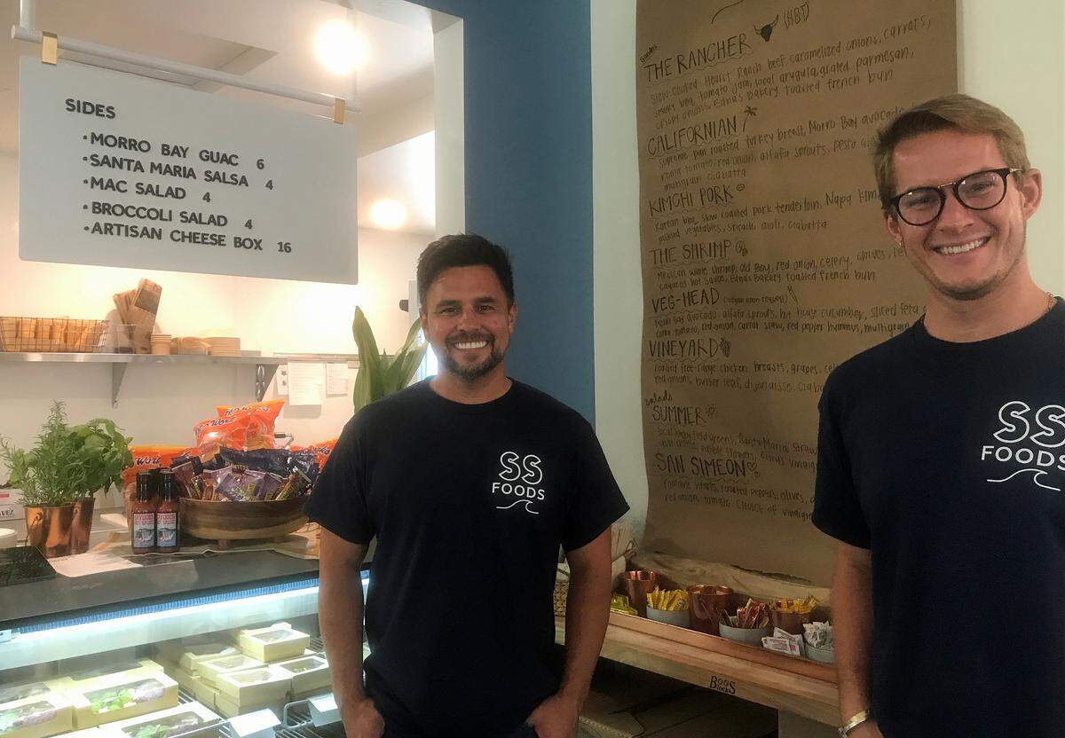 Alex Labastida, at left, and husband Alex Saleen pose for a picture by the wall-mounted menu for Seaside Foods deli inside the recently reopened Sebastian’s General Store in Old San Simeon Village on Friday, June 16, 2023.