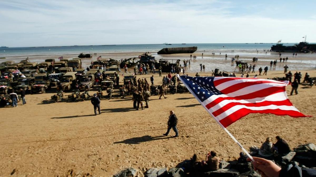 An onlooker waves an American flag as World War II military vehicles are displayed on the beach of Arromanches, France, Friday, June 6, 2014, as part of D-Day commemorations. World leaders and veterans gathered by the beaches of Normandy, northern France, to mark the anniversary of the World War II D-Day landings.